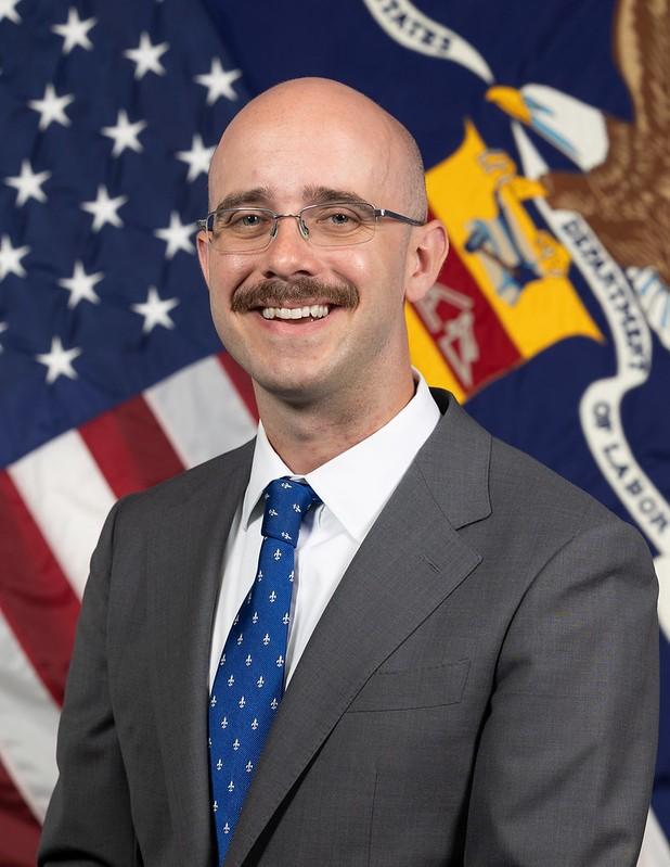 A bald man wearing a suit and glasses smiles while seated in front of the U.S. and Labor Department flags.