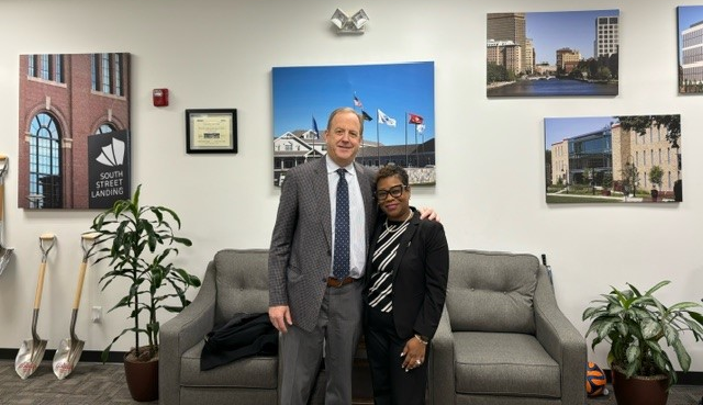 A man and woman in business clothes stand in front of a wall with photos showing completed construction projects.