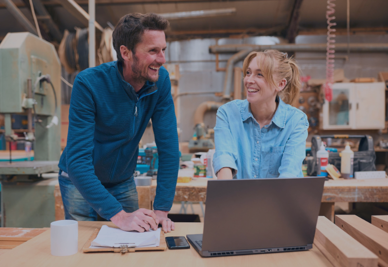 A man and a woman in a workshop are laughing and talking while looking at something on a laptop.