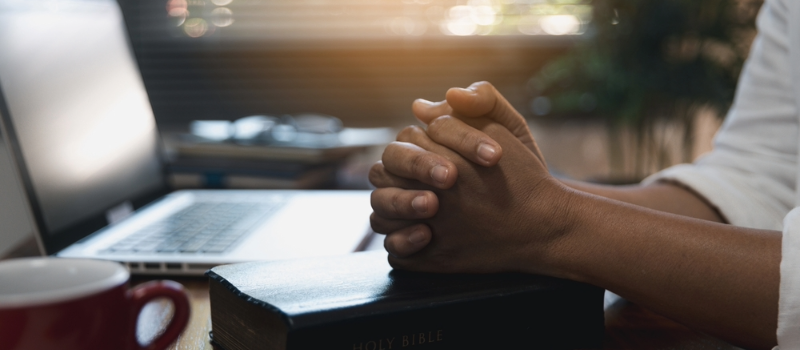 Close-up of a desktop with a laptop, mug and bible. A person sits with their hands clasped over the bible.