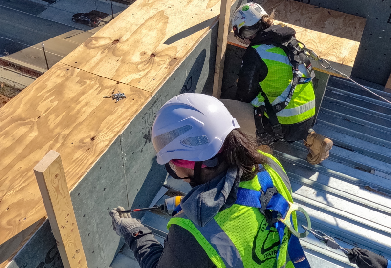 Two female construction employees wearing fall protection gear, including safety helmets, harnesses and other equipment.