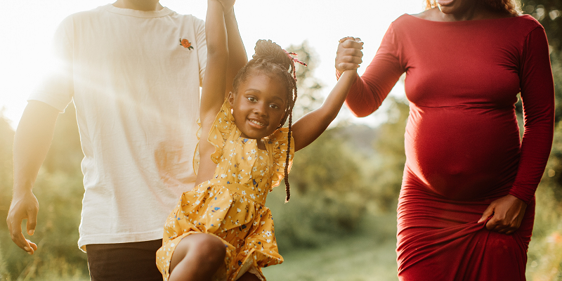A Black family holds hands: a dad, a young daughter and a pregnant mother.