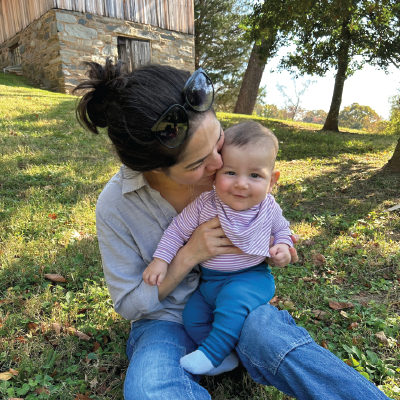 A mother sits on a lawn, holding a smiling baby in her lap and giving it a gentle kiss.