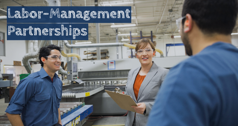 A supervisor holding a clipboard speaks with two workers in a factory environment. They all wear safety goggles and ear plugs. The text reads "Labor-Management Partnerships."