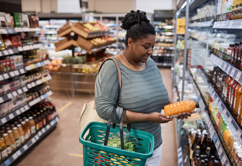 A woman holding a shopping basket looks at a bottle of juice in a grocery store refrigerator aisle. 