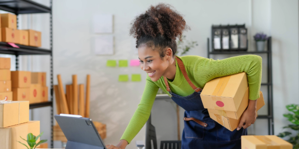 A young entrepreneur checks her laptop while holding shipping boxes.