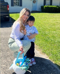 A woman crouches in a driveway to embrace a young boy. She holds an Easter basket full of colorful eggs.