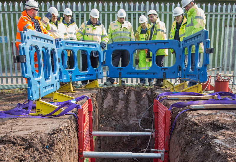 A group of workers in high visibility clothing and hard hats stand around a trench that has protective barriers preventing them from falling in as well as shoring inside to keep it from collapsing.