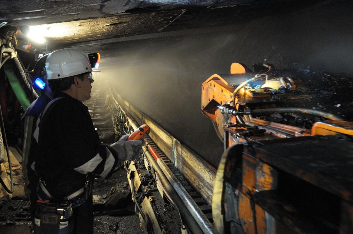 A coal miner wearing a helmet with a headlamp controls a continuous mining machine underground.