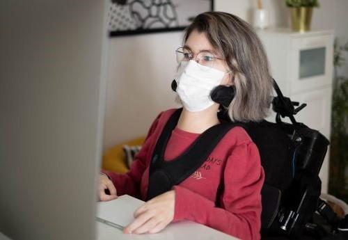 Woman wearing a mask, sitting in a wheelchair, and working on a computer