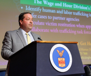 The image shows a person standing at a podium with the U.S. Department of Labor seal on it. The person is dressed in a suit and is delivering a speech. A presentation slide is visible in the background, related to human and labor trafficking topics.