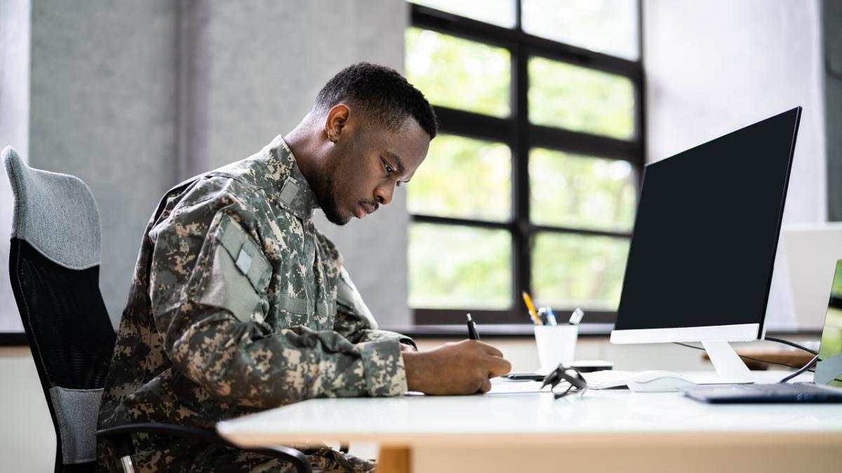 African American veteran in military uniform writing at a desk beside a computer in an office