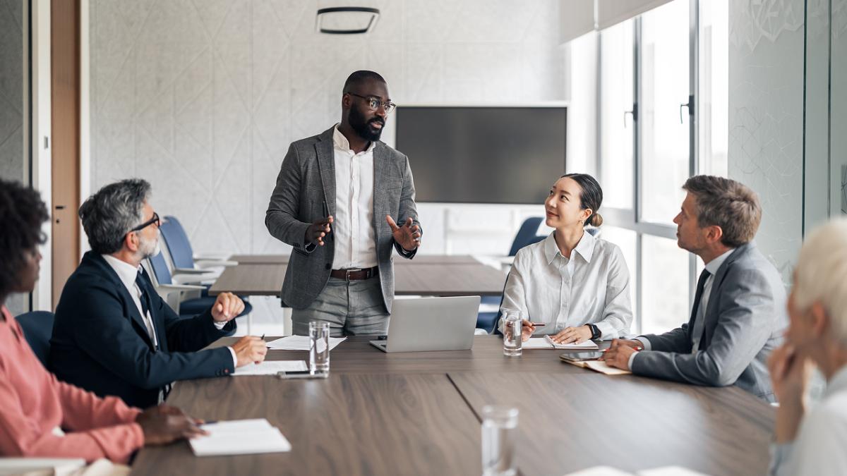 Black businessman presenting strategy to a diverse team in a modern boardroom, colleagues listening attentively around a conference table with laptop and documents