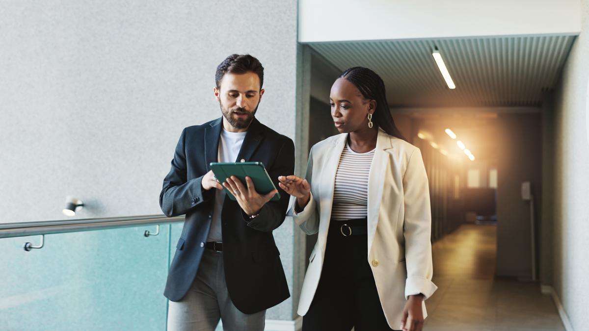 Two business professionals walk through an office hallway while reviewing information on a tablet together