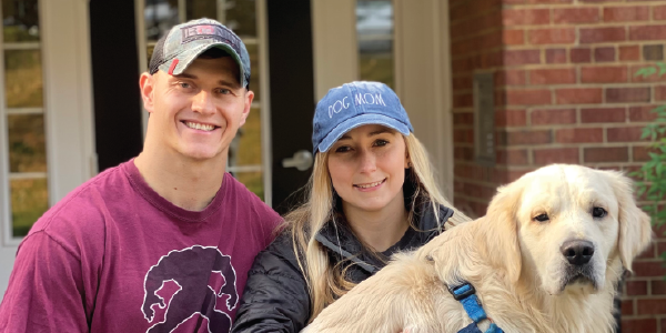 A young couple poses in front of a home. The woman is holding a large vanilla lab.