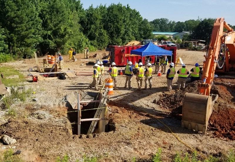 View of a trench at a construction site  with an excavator and workers walking around 