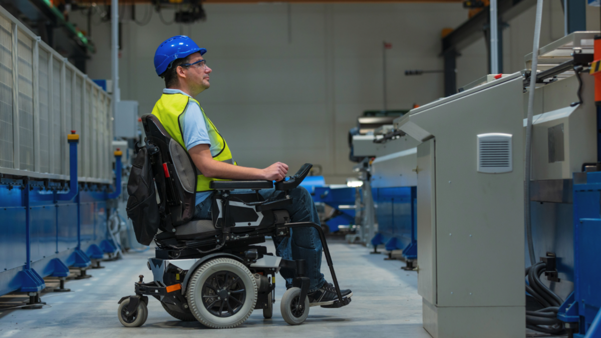 A worker in a powered wheelchair operates machinery inside a large industrial facility. The person wears a hard hat and a high‑visibility safety vest while positioned at a control panel between rows of manufacturing equipment.