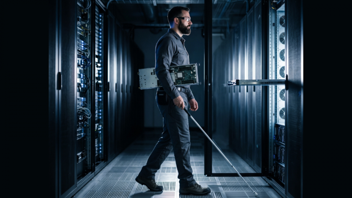 An information technologist navigates a data center using a white cane while carrying computer hardware. Rows of illuminated server racks line both sides of the corridor with a door open.